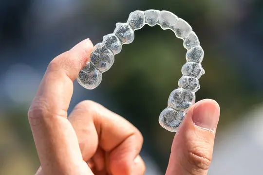 A hand holds a clear Invisalign aligner. Chauncey Orthodontics in Henderson, NV offers these nearly invisible teeth straightening solutions.