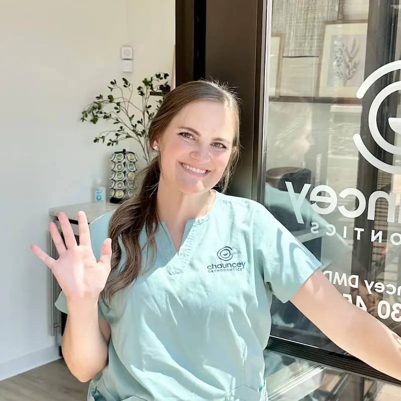 A smiling woman in scrubs with the Chauncey Orthodontics logo waves. The clinic is located in Henderson, NV.