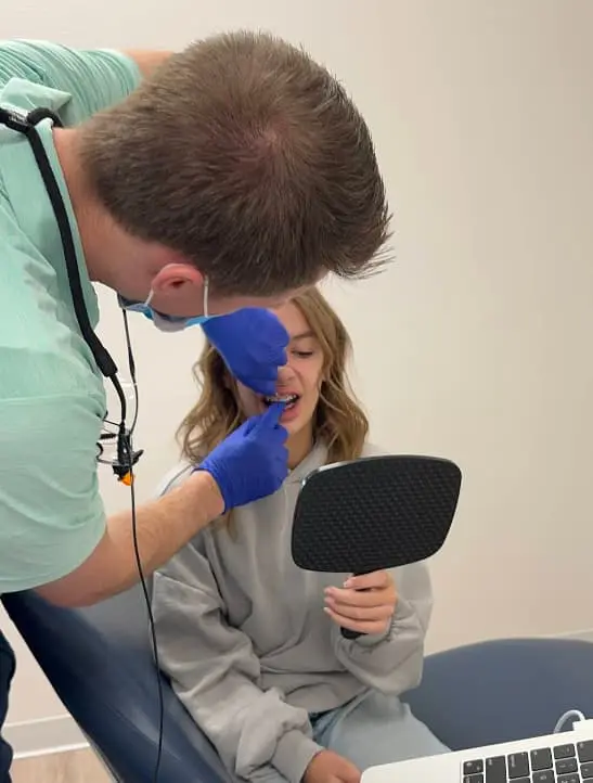 An orthodontist examines a young patient's teeth while the patient holds a mirror. Chauncey Orthodontics in Henderson, NV, provides orthodontic care.