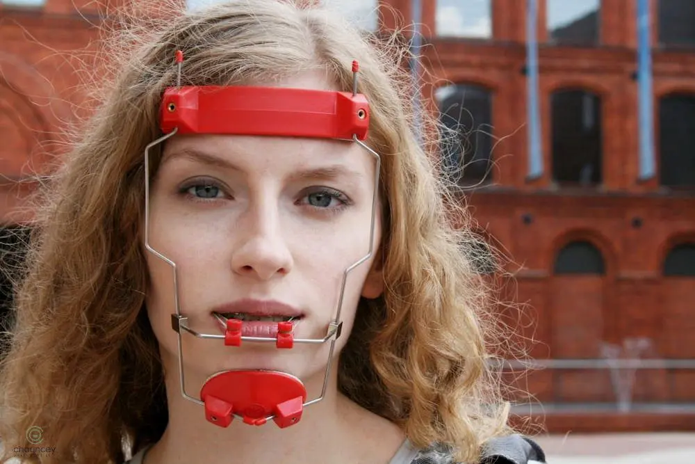 a woman looking at the camera wearing red reverse pull headgear braces in Henderson, NV A person with curly blonde hair wears a red and metal orthodontic headgear on their face, standing in front of a red brick building in Henderson, NV.