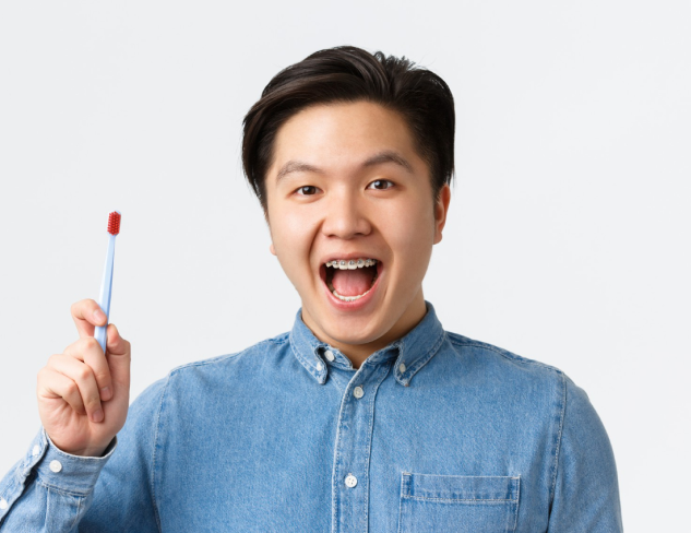 A person in a denim shirt, highlighting their braces while holding a toothbrush, represents adult orthodontic care with Chauncey Orthodontics in Henderson, NV set against a simple light background.