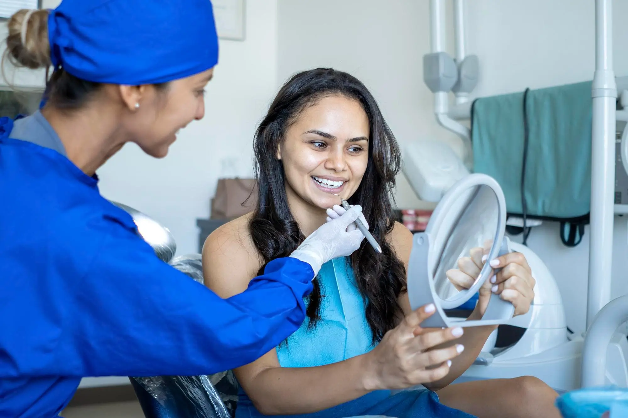 A dental professional in blue scrubs shows dental tools to a smiling woman holding a mirror at a dental clinic with discuss adult orthodontics with a patient at Chauncey Orthodontics in Henderson, NV.