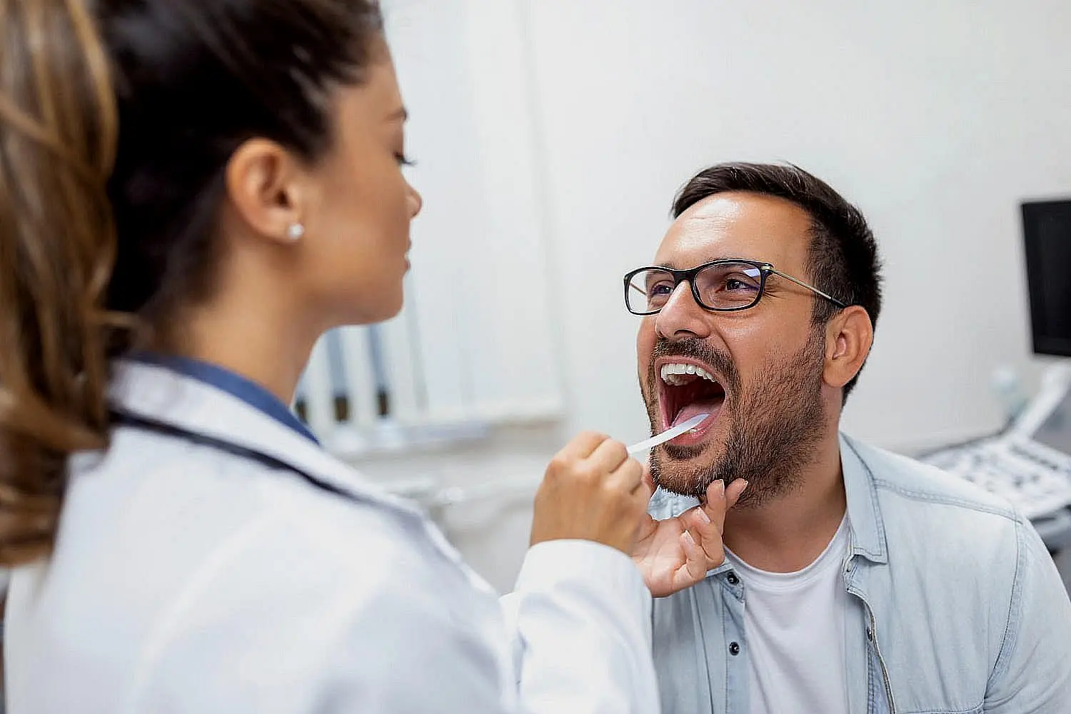A healthcare professional uses a tongue depressor to examine the open mouth of a man wearing glasses during therapy a tongue thrust at Chauncey Orthodontics operates in Henderson, NV.