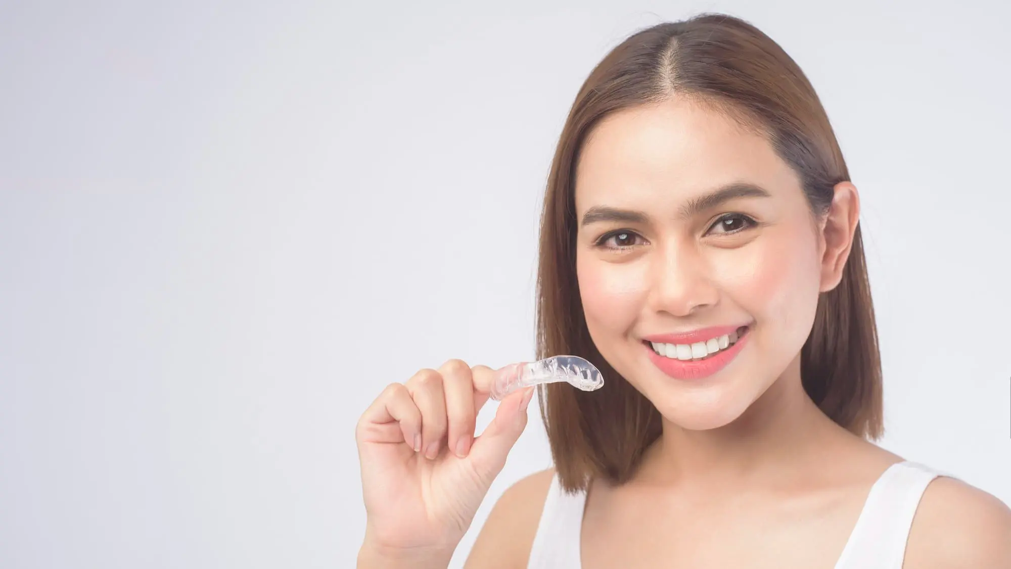 A woman with straight brown hair smiles while holding a clear dental aligner in her right hand against a plain white background.