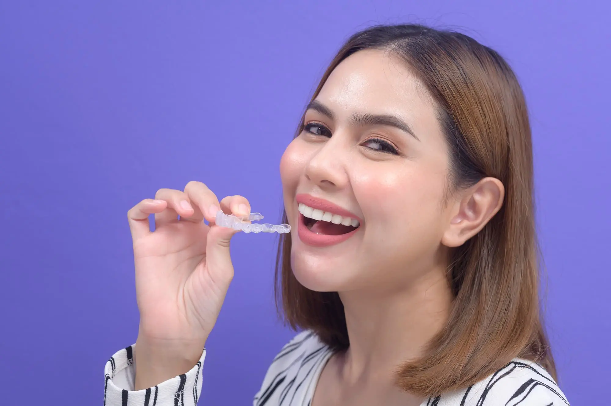 A woman smiles while holding a clear dental aligner near her mouth against a purple background.