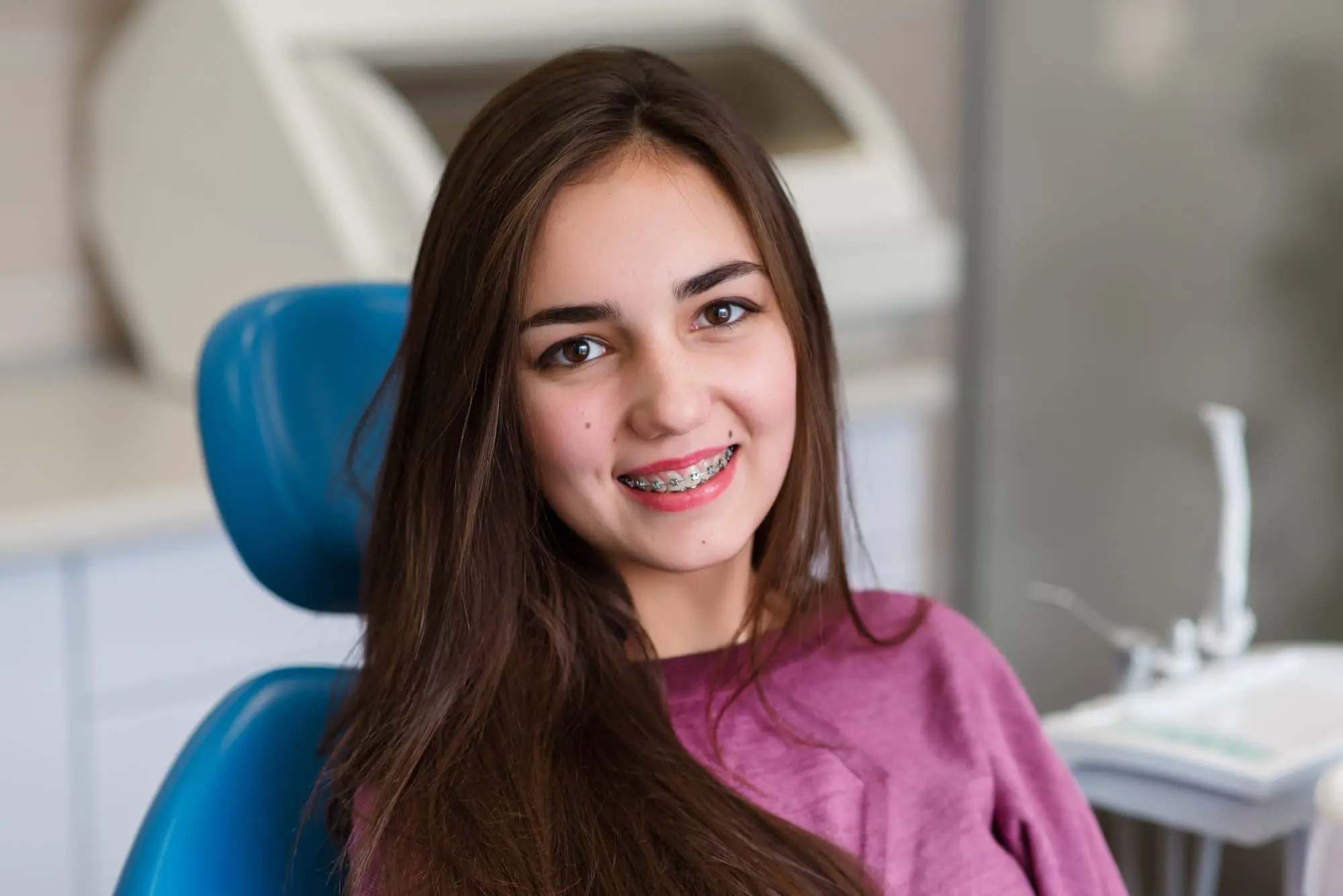 A young woman with long brown hair and braces sits in a dental chair, smiling at the camera in a dental clinic setting.