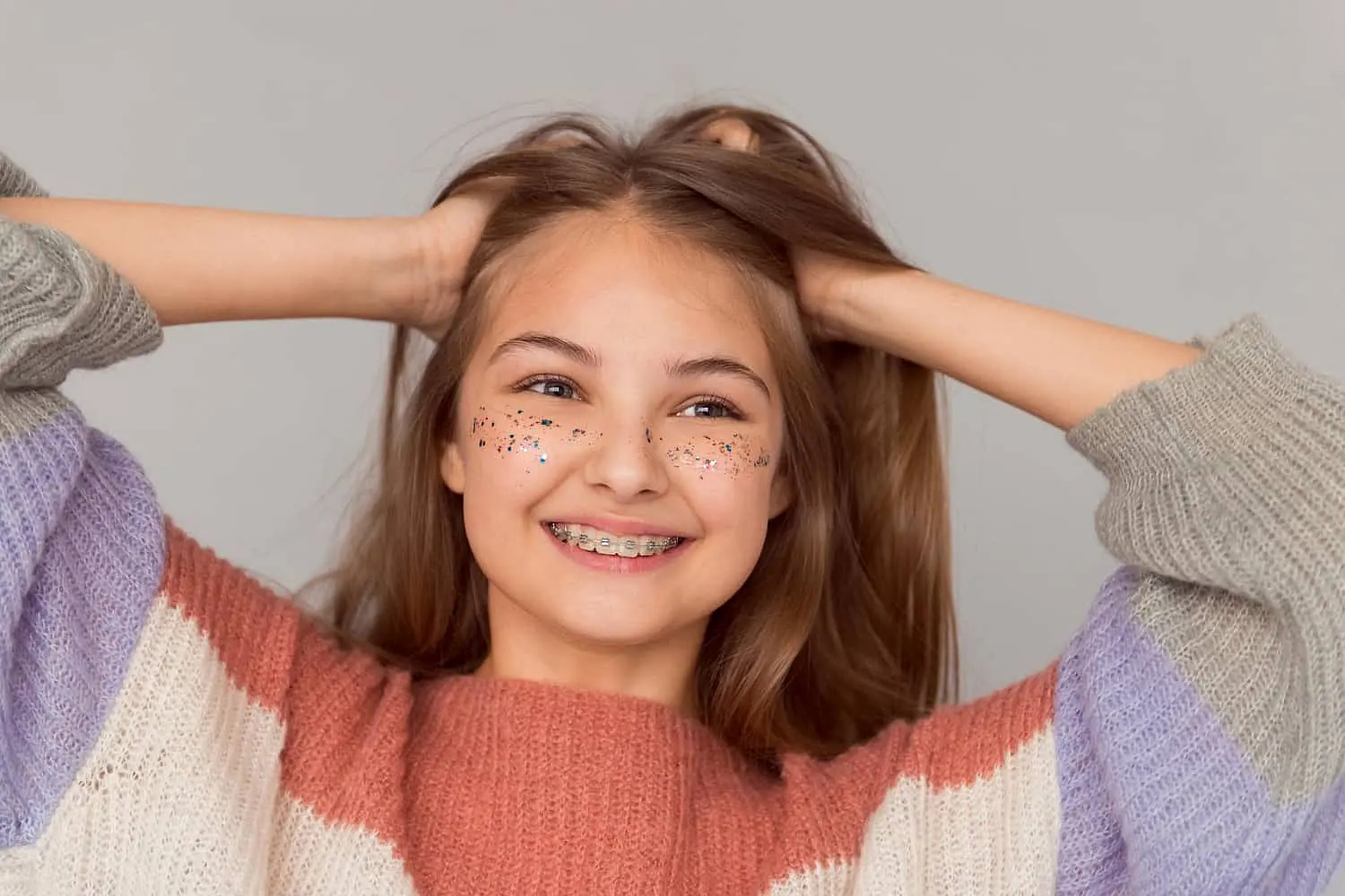 A smiling girl with traditional braces and glittery cheeks, dressed in a striped sweater, confidently holds her hair back against a plain background showcasing the positive results achieved at Chauncey Orthodontics in Henderson, NV.