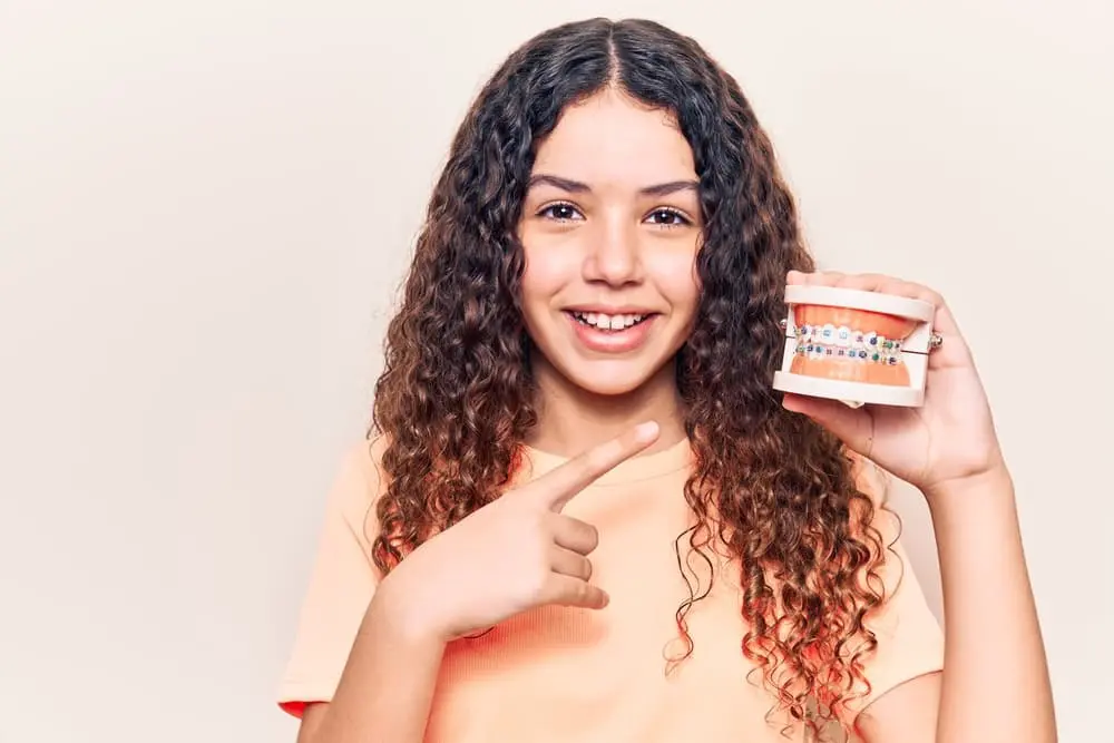 Best Orthodontics A smiling young person with curly brown hair points to a model of teeth with braces. Contact Chauncey Orthodontics in Henderson, NV, for braces cost information.