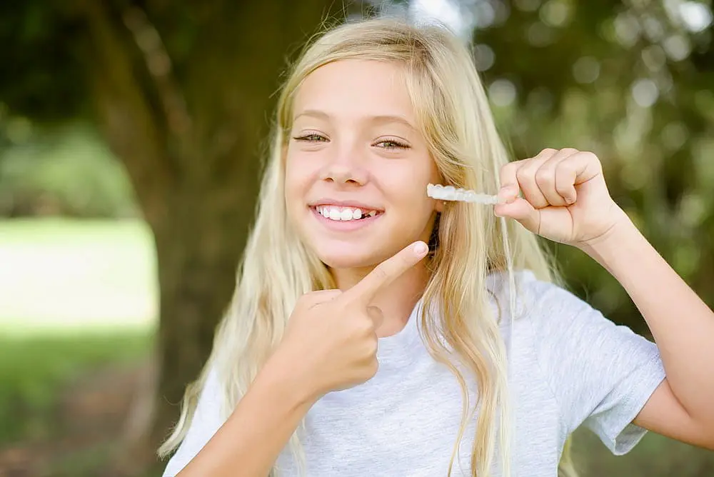 A smiling girl with long blonde hair stands outdoors, holding and pointing to a clear aligner, highlighting the benefits of visiting Chauncey Orthodontics in Henderson, NV for a children's orthodontist.