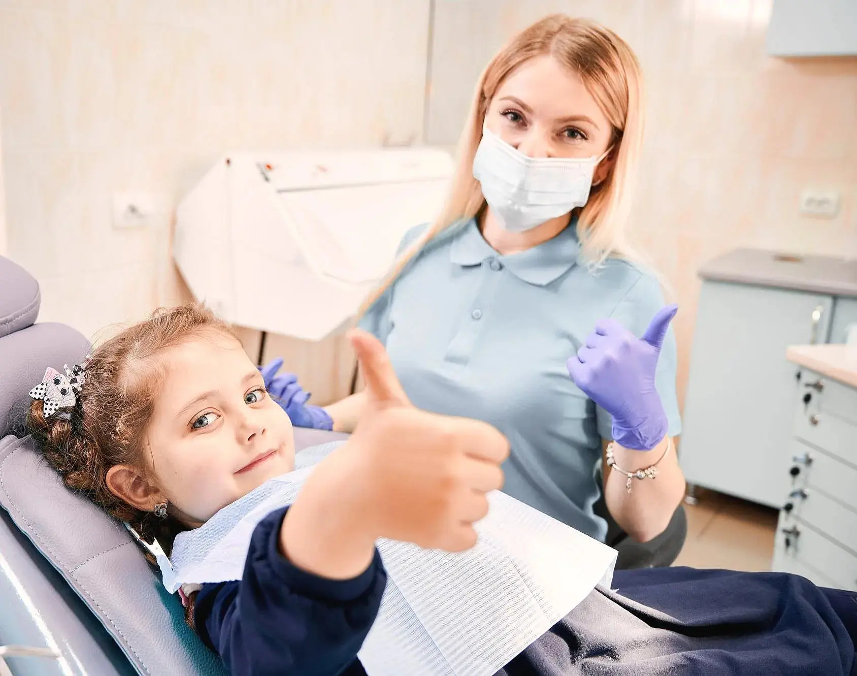 A young girl sits in a dental chair giving a thumbs up, while a children orthodontist wearing gloves and a mask also gives a thumbs up in Henderson, NV.