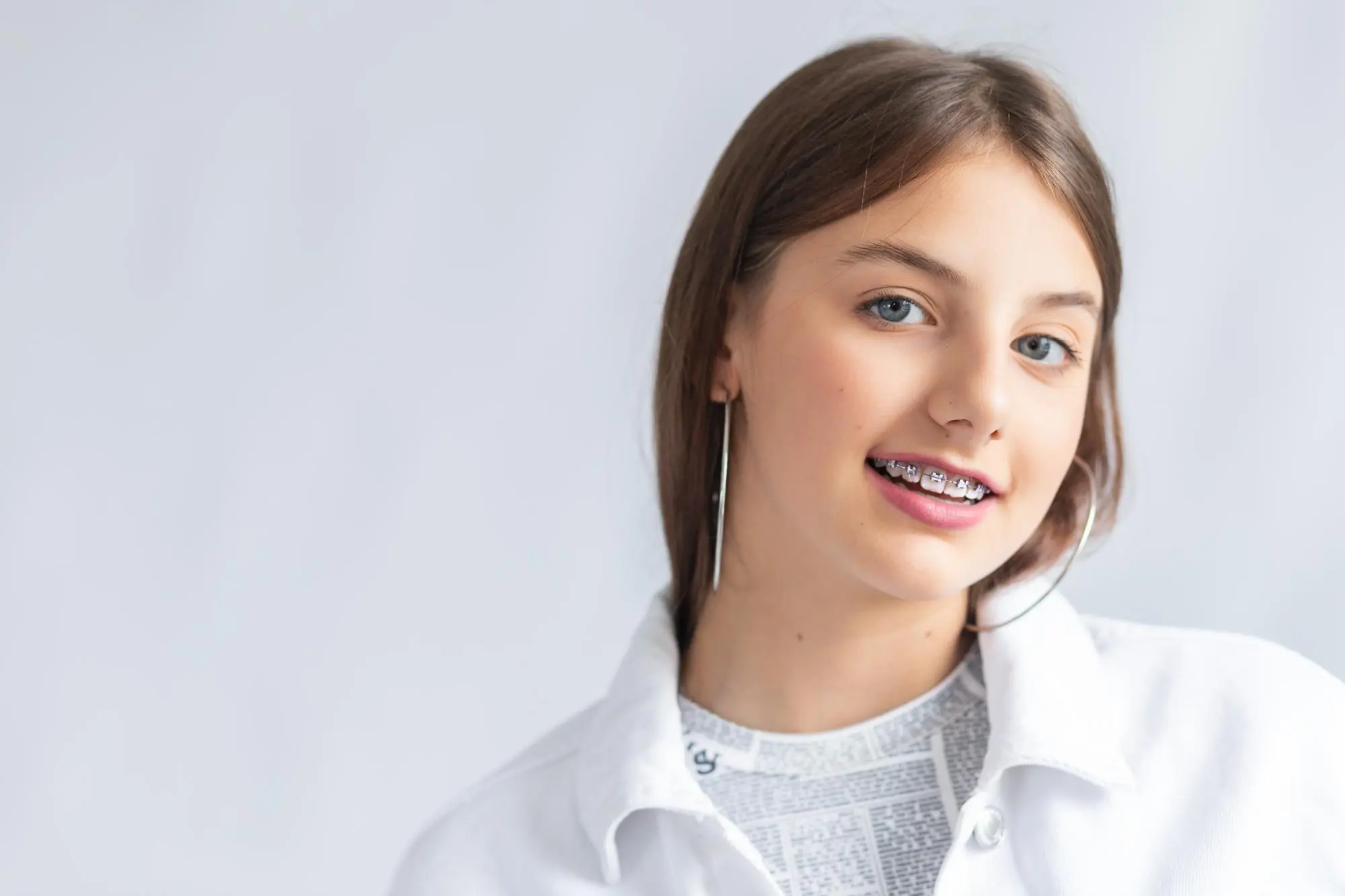 A young person with long brown hair and braces smiles at the camera, wearing a white shirt and hoop earrings against a light background.
