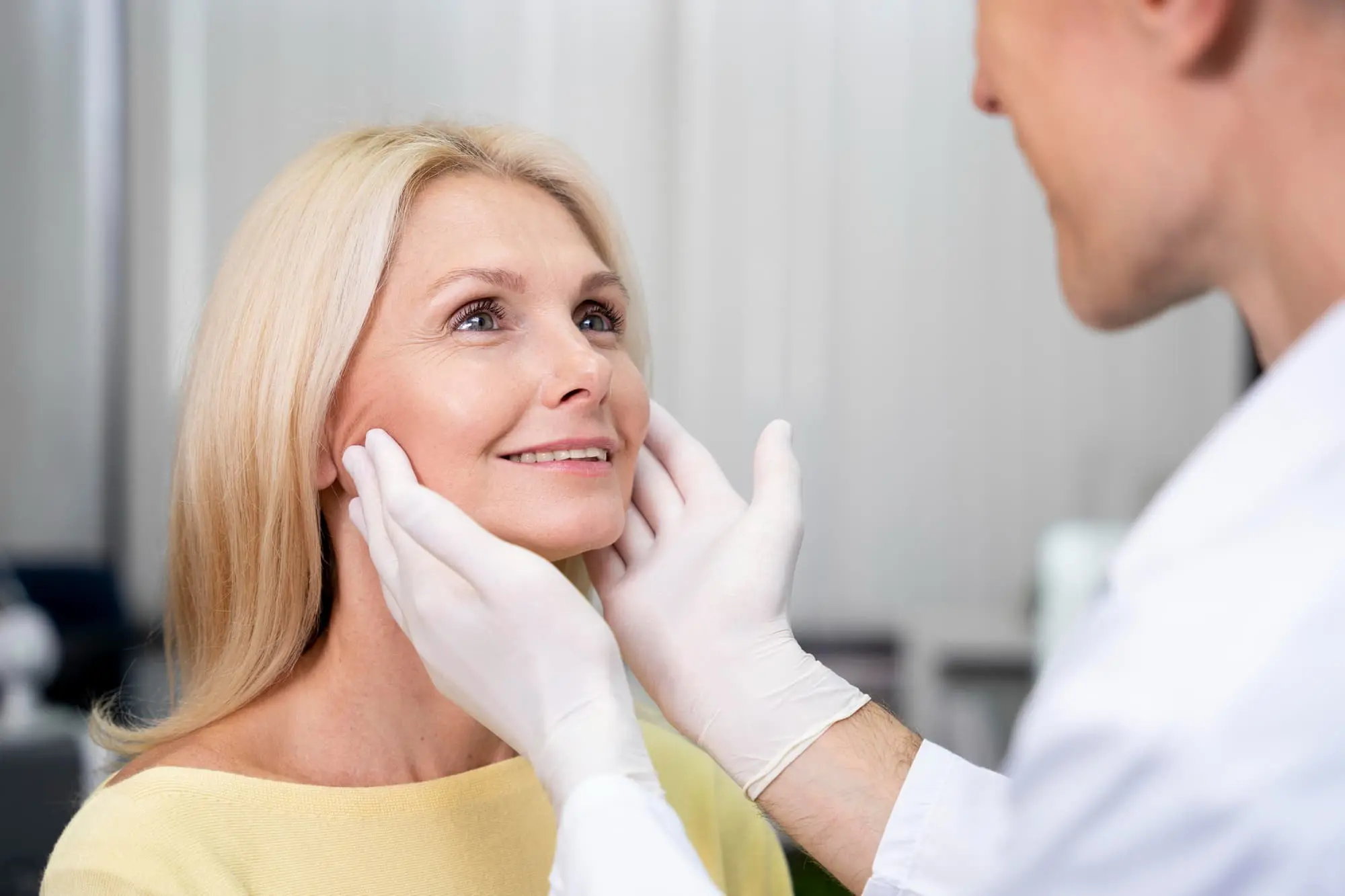 A doctor wearing gloves examines a middle-aged woman's face as she smiles in a medical office.