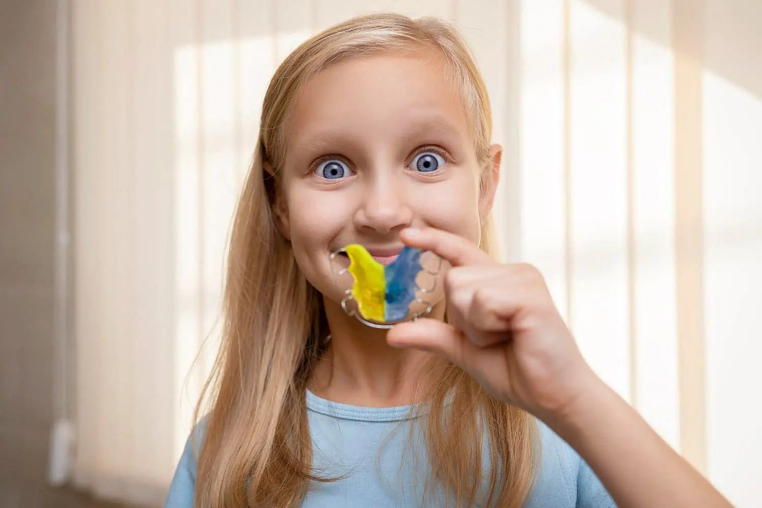 A young girl with long blonde hair smiles at the camera while holding a blue and yellow dental retainer, highlighting her visit to Chauncey Orthodontics in Henderson, NV.