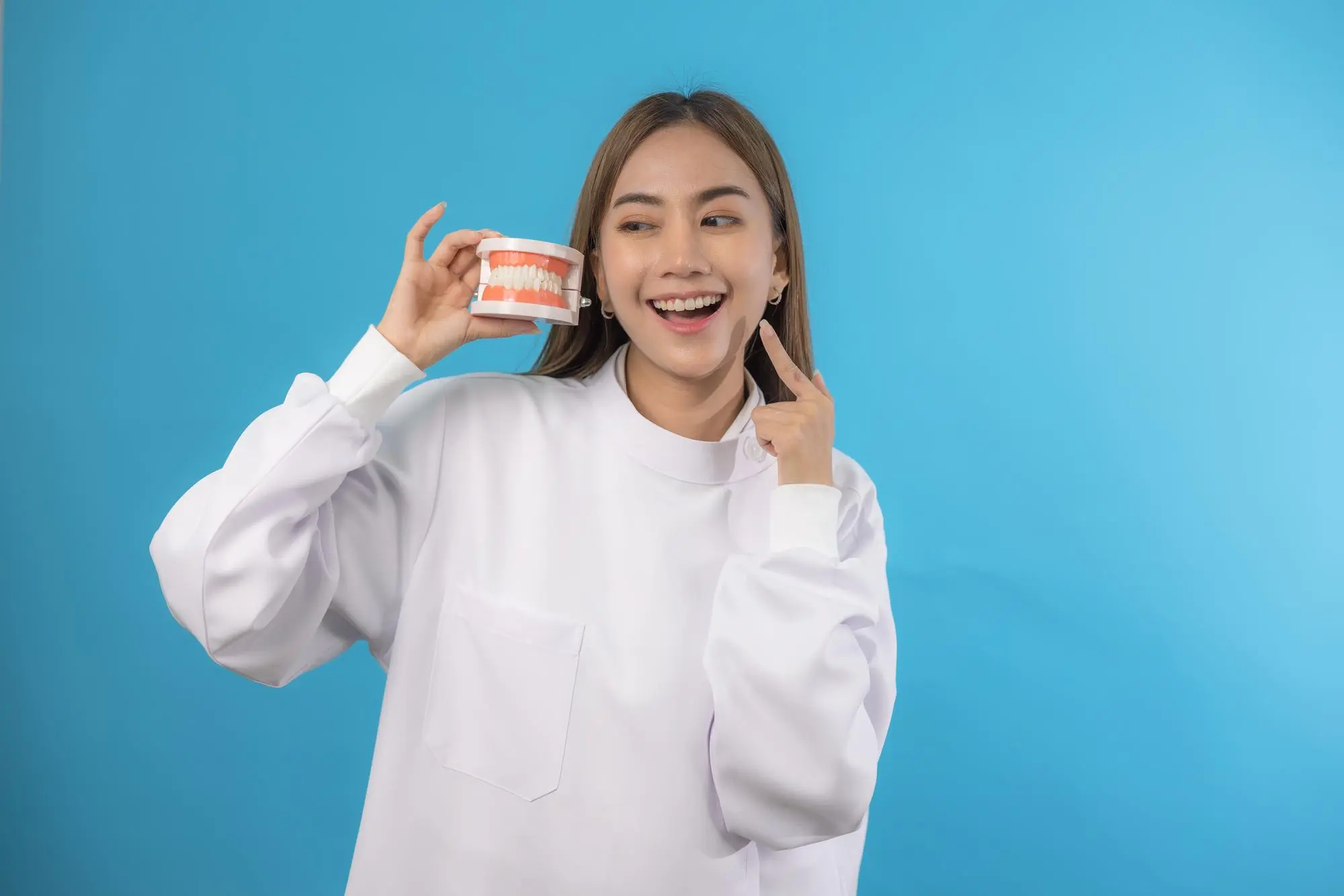 A woman in a white shirt holds a dental model next to her face and points to her mouth, standing against a blue background.