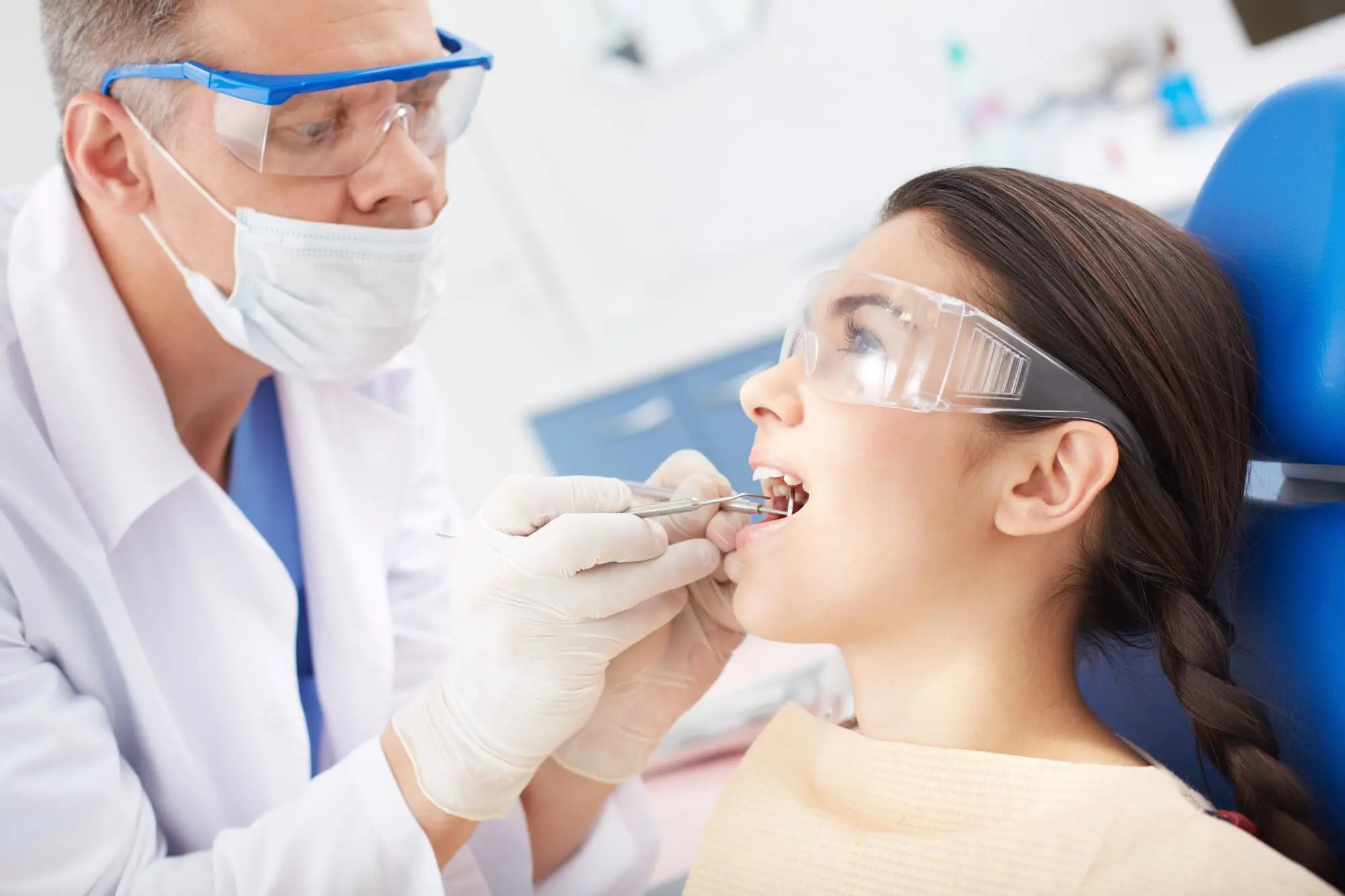 A dentist wearing a mask and safety glasses examines a patient's teeth while the patient sits in a dental chair wearing protective eyewear.