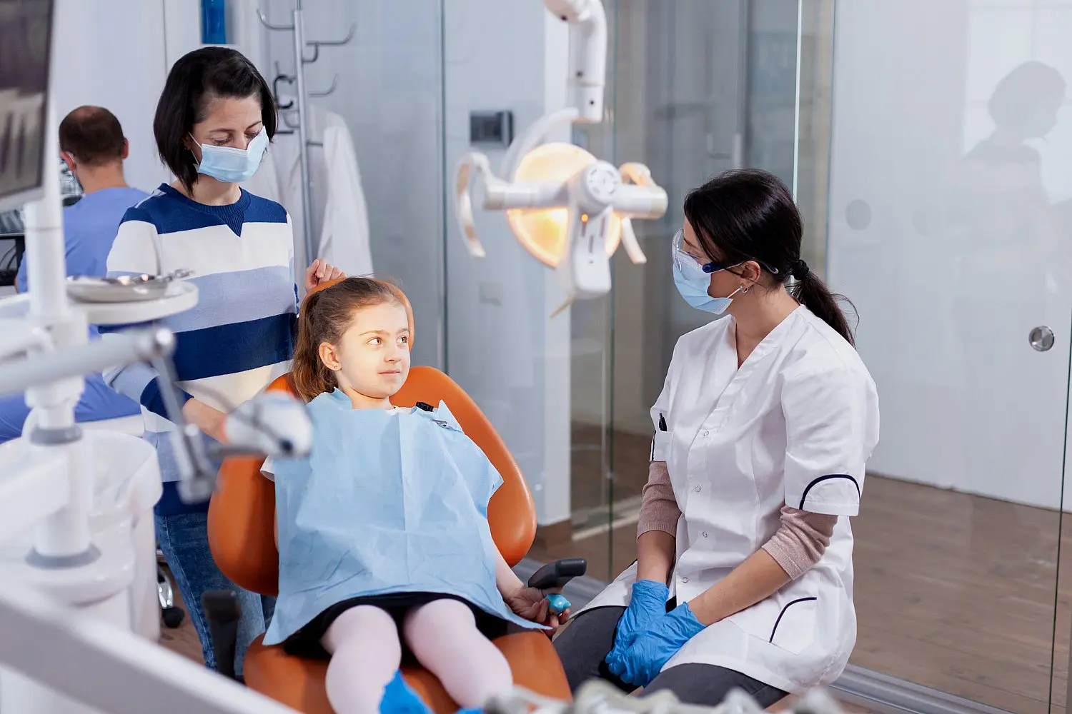 At Chauncey Orthodontics in Henderson NV, a children’s orthodontist in a white coat and gloves warmly speaks to a young girl sitting with an adult in the dental chair.