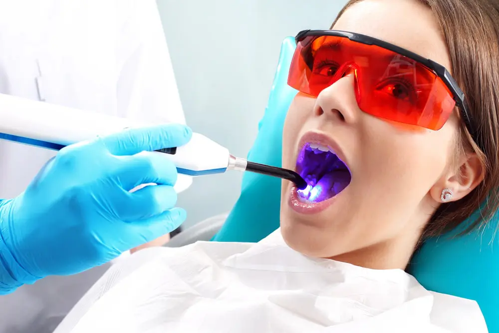 A person at Chauncey Orthodontics operates in Henderson, NV receives dental treatment, comparing invisalign vs braces, while using a blue light device with protective red glasses and a dental bib.