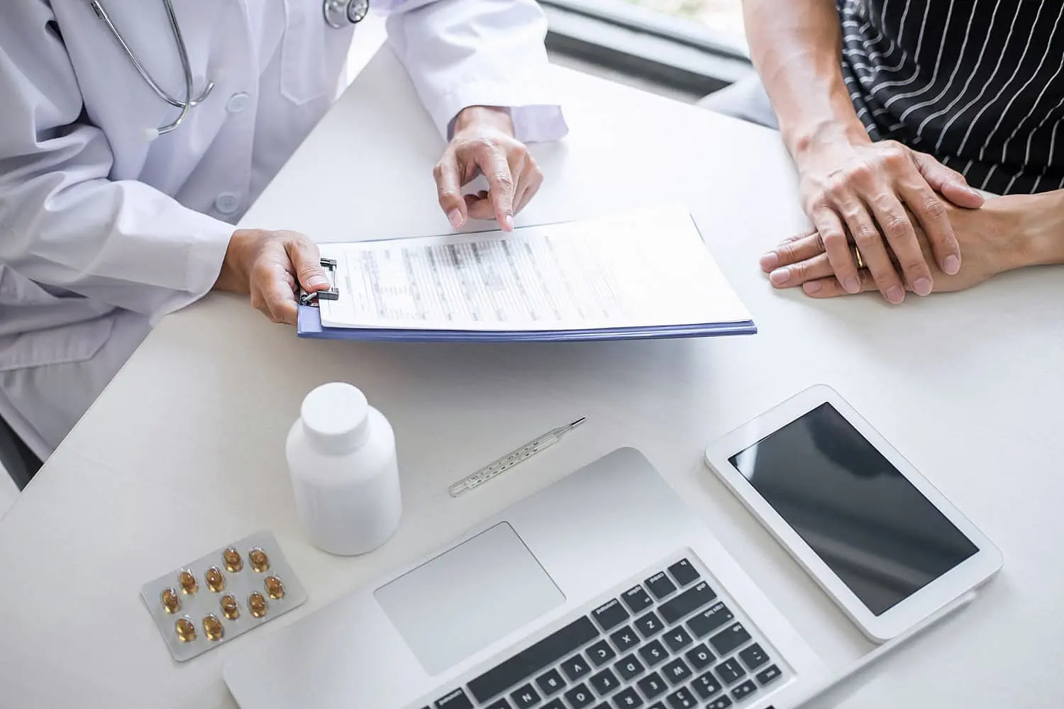 A doctor discusses orthodontic insurance plans with a patient, showing a clipboard with documents across a desk adorned with a laptop, tablet, pills, and a bottle in Henderson, NV.