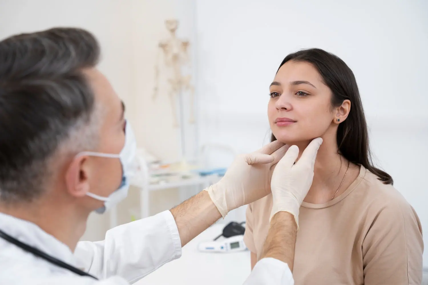A doctor in a mask checks the throat of a seated female patient, discussing potential jaw surgery options in a modern medicine office in Henderson, NV.
