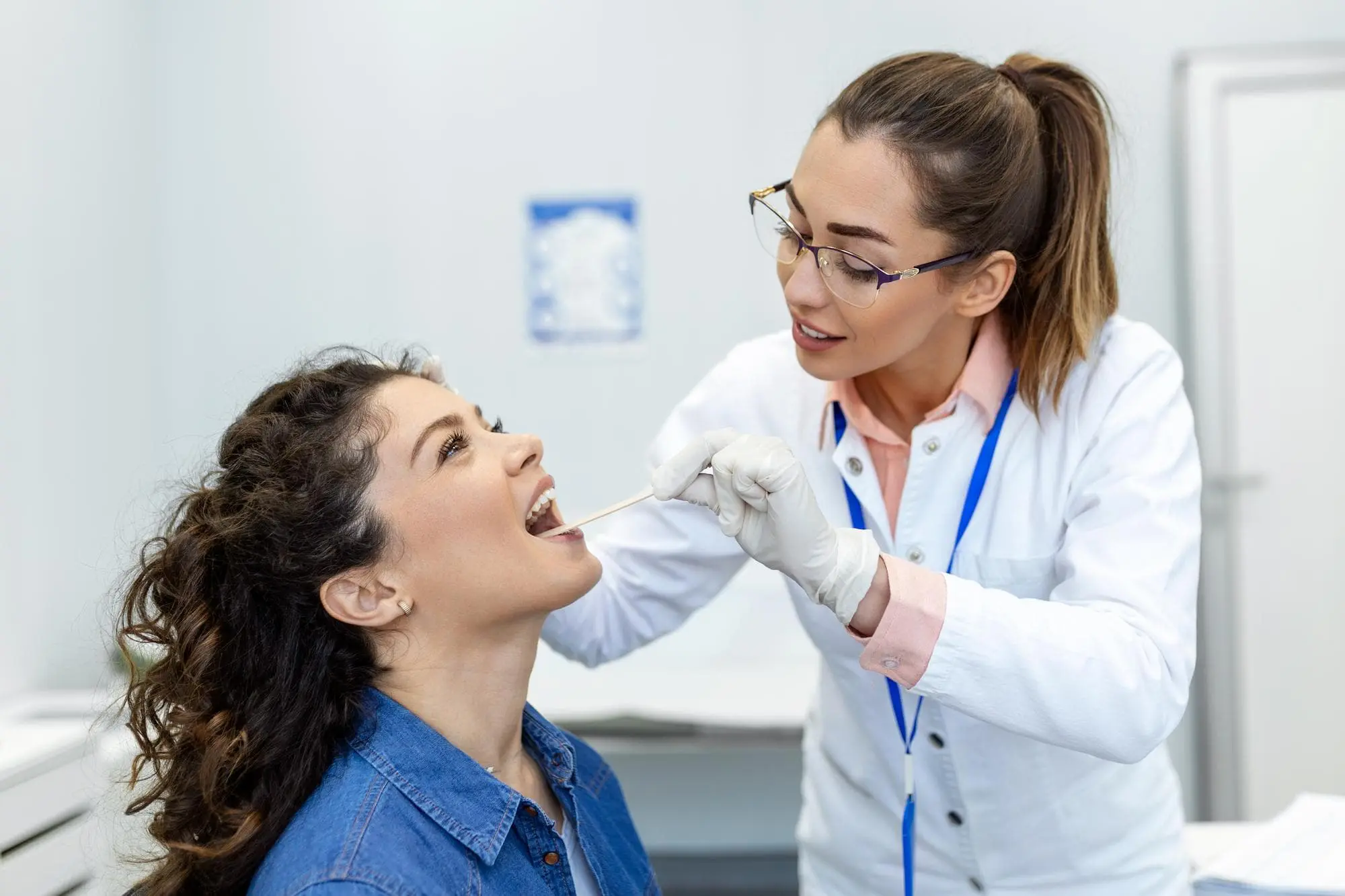 A healthcare professional in a white coat uses a tongue depressor to examine a seated patient's open mouth in a medical office.