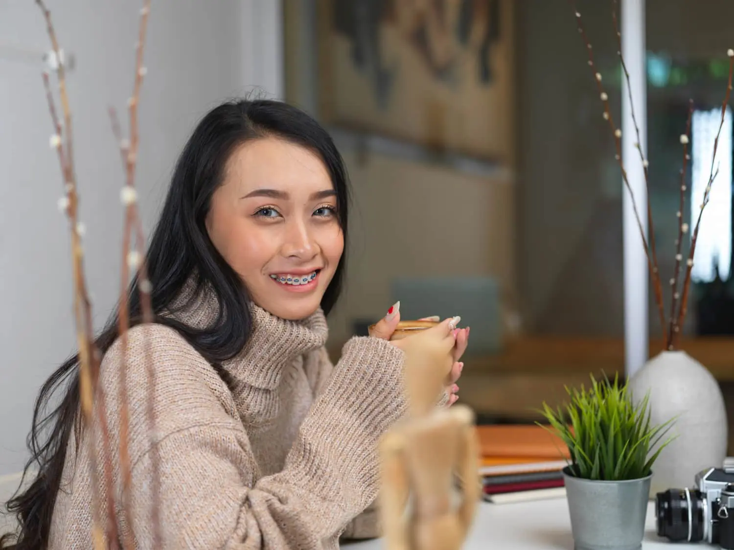 A woman with long dark hair and braces, wearing a beige sweater, sits at a table holding a mug. There are plants and notebooks on the table.