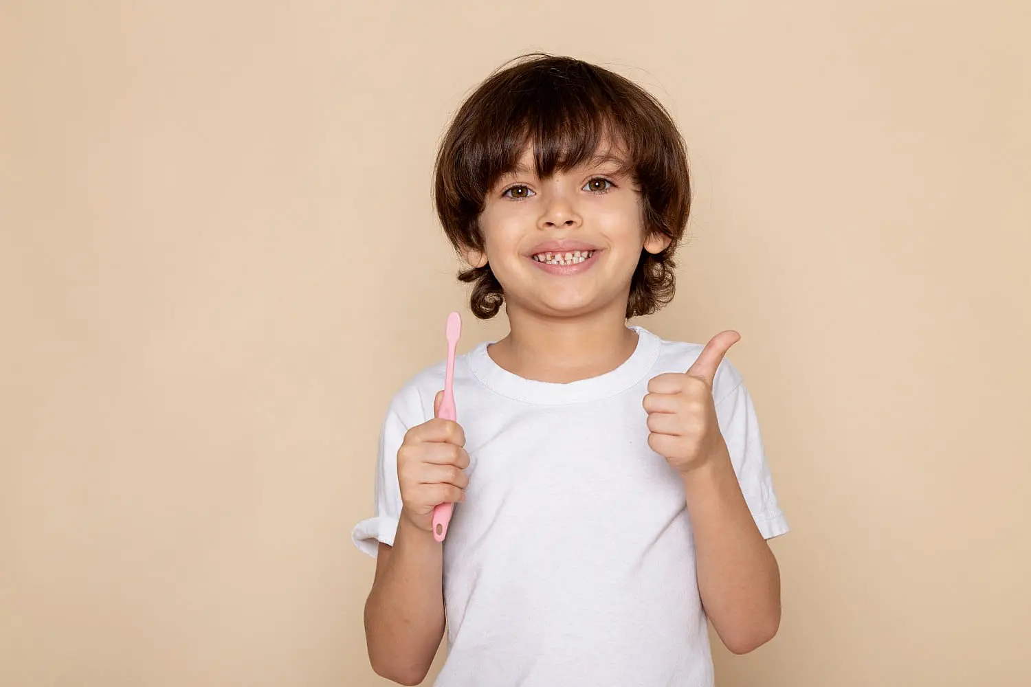 A young child in a white t-shirt, with braces and a bright smile, holds a pink toothbrush and gives a thumbs-up after visiting children orthodontist at Chauncey Orthodontics in Henderson, NV.