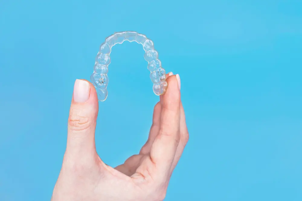 A hand holds a clear Invisalign aligner against a blue background, suggest choose affordable invisalign with invisalign cost at Chauncey Orthodontics in Henderson, NV.