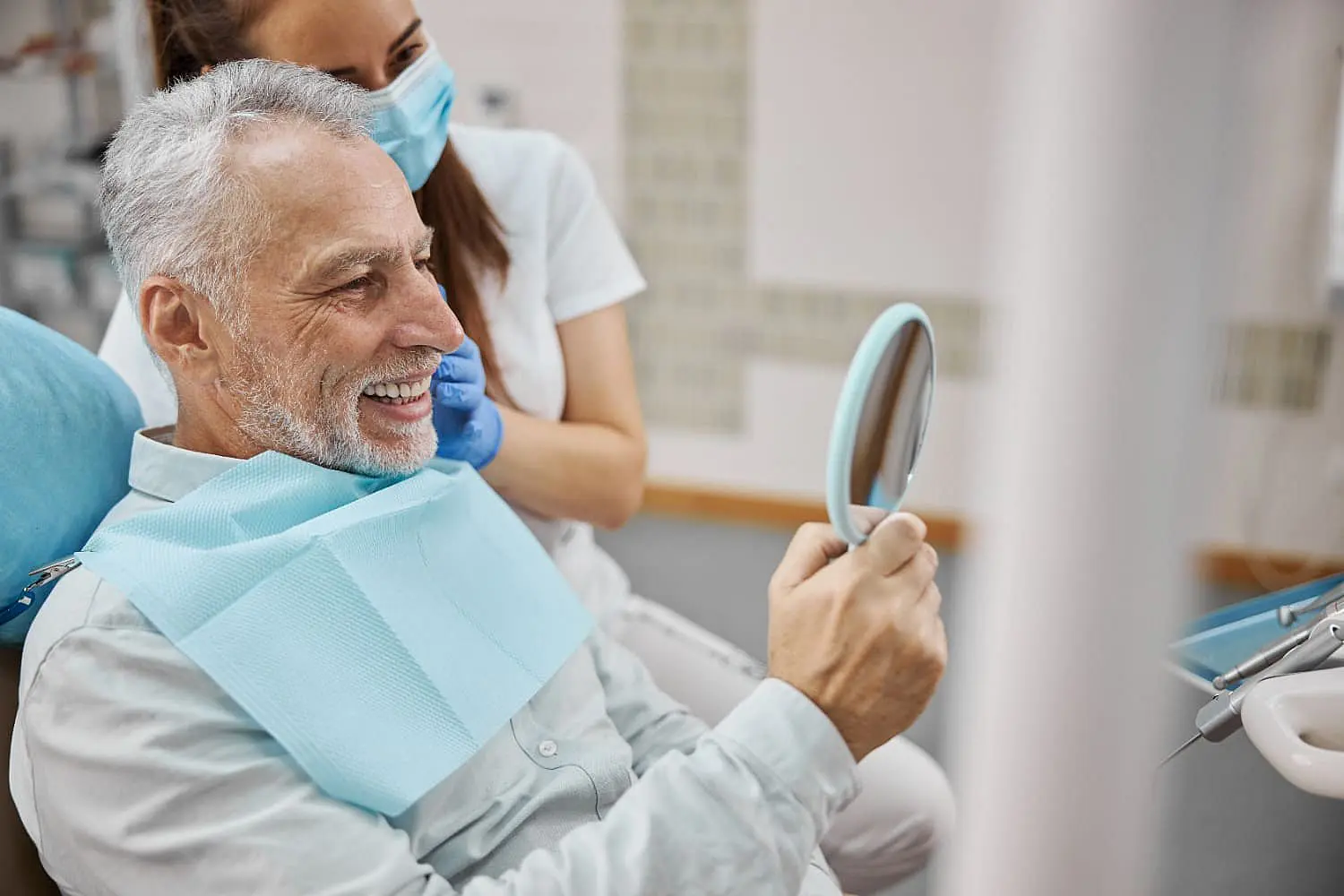 An older man sits in a dental chair, smiling as he looks into a handheld mirror happy with adult orthodontics from Chauncey Orthodontics in Henderson, NV.