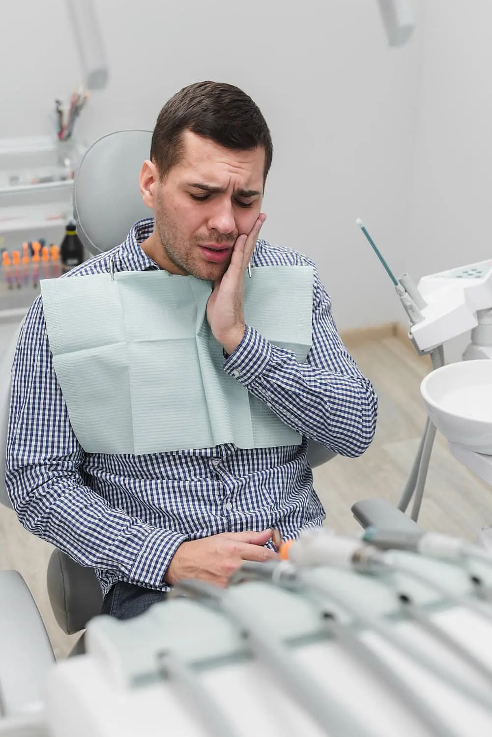 In a dental chair, a man holds his cheek in discomfort, possibly contemplating the Invisalign vs Braces debate, surrounded by dental tools. For those facing similar choices, Chauncey Orthodontics in Henderson, NV can help.