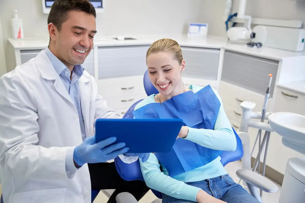 A dentist wearing gloves shows a tablet to a smiling patient sitting in a dental chair in a modern clinic.