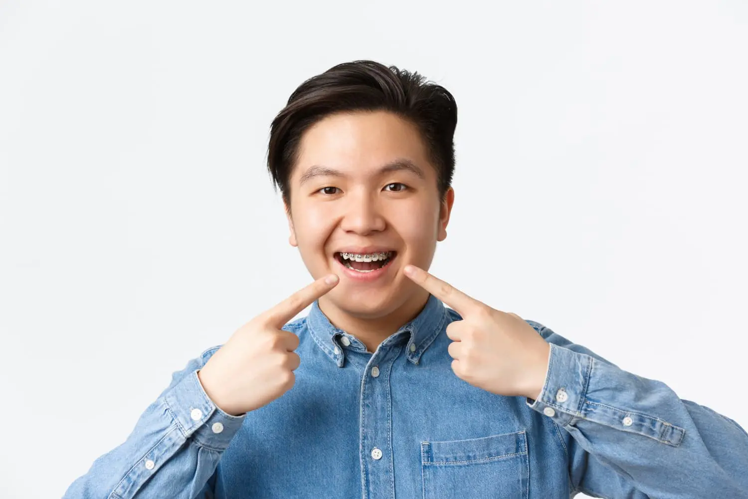 A person wearing a denim shirt beams with confidence, proudly showcasing their adult braces against a plain white background in Henderson, NV.