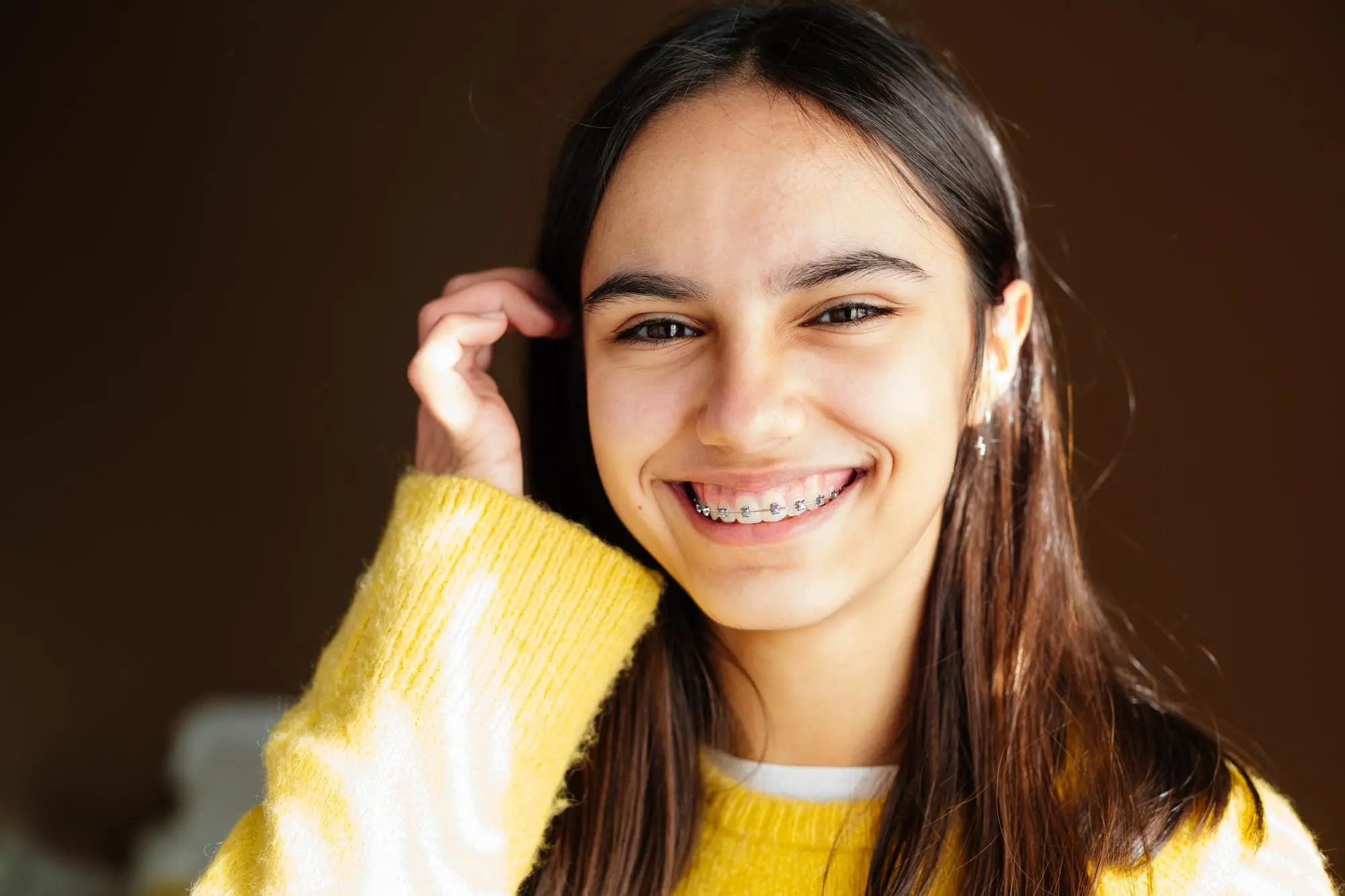 A young person with long dark hair, wearing a yellow sweater and braces, smiles at the camera against a plain brown background.