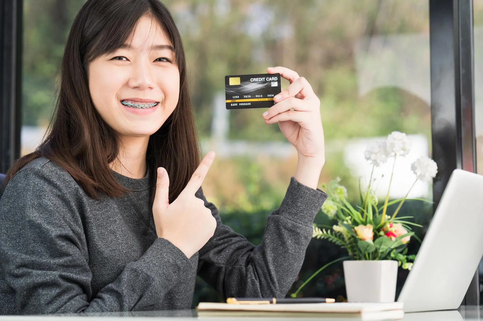 A young woman with braces smiles and points at a credit card she is holding, seated at a desk with a laptop, notebook, and a potted plant.