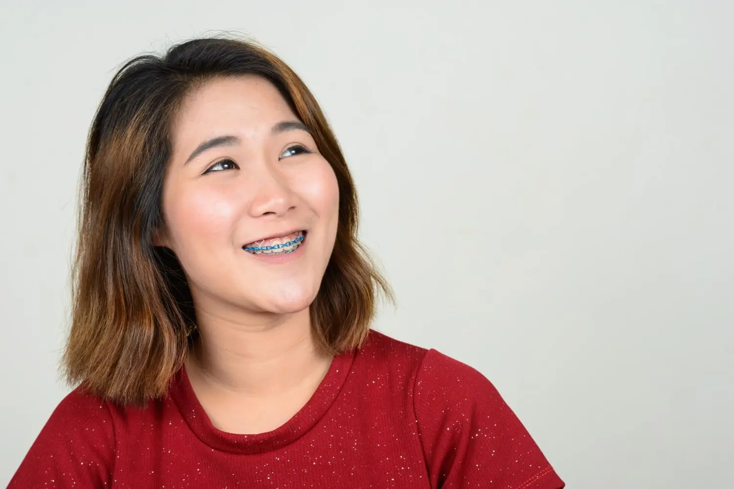 At Chauncey Orthodontics in Henderson NV, a young woman with shoulder-length hair and adult braces smiles to her left, dressed in a red shirt against a plain light background.