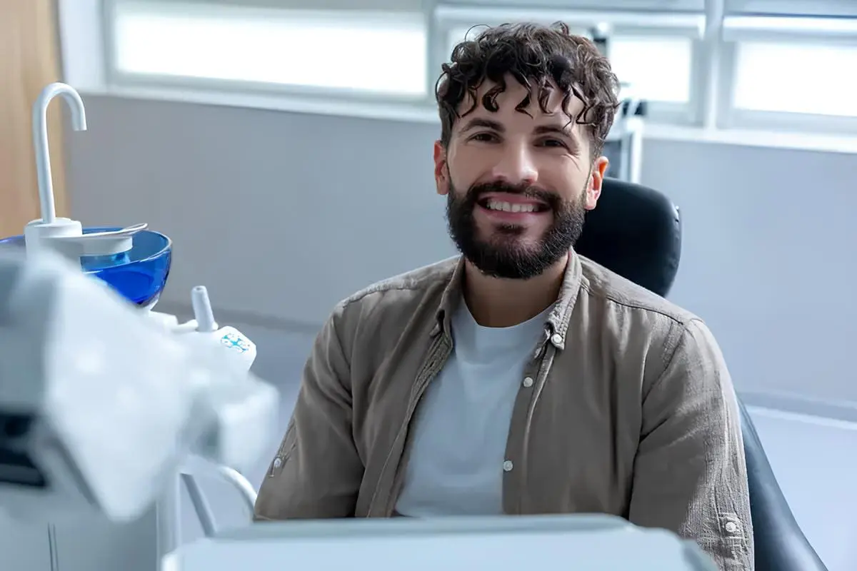 A man with a beard sits and smiles in a dental chair in a brightly lit clinic. Dental equipment is visible in the foreground.