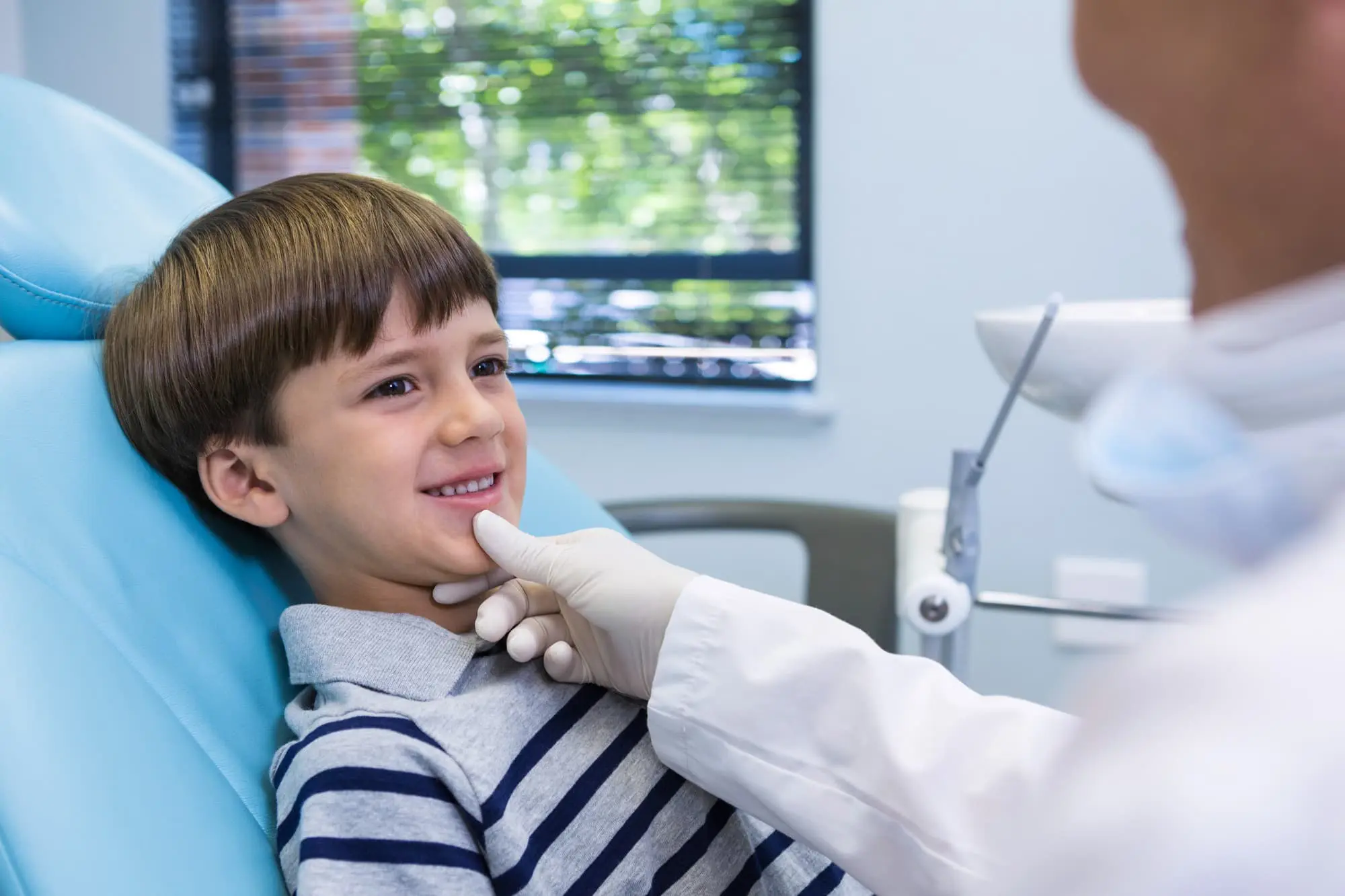 A young boy sits in a dental chair, smiling, while a dentist wearing a white coat and gloves gently touches his chin during a check-up with children orthodontist at Chauncey Orthodontics in Henderson, NV.