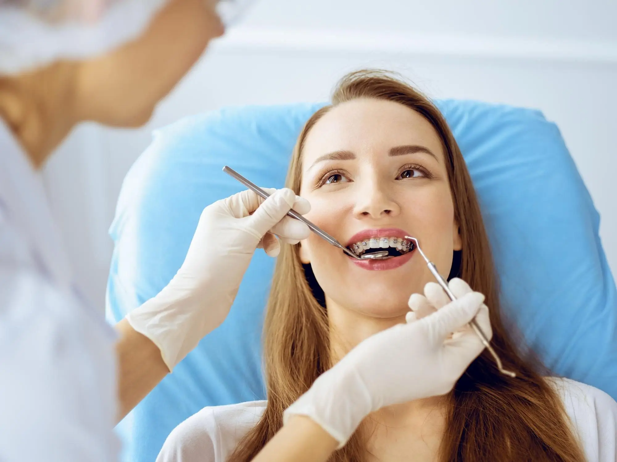 A dentist examines a young woman with braces, using dental tools while she sits in a blue chair.