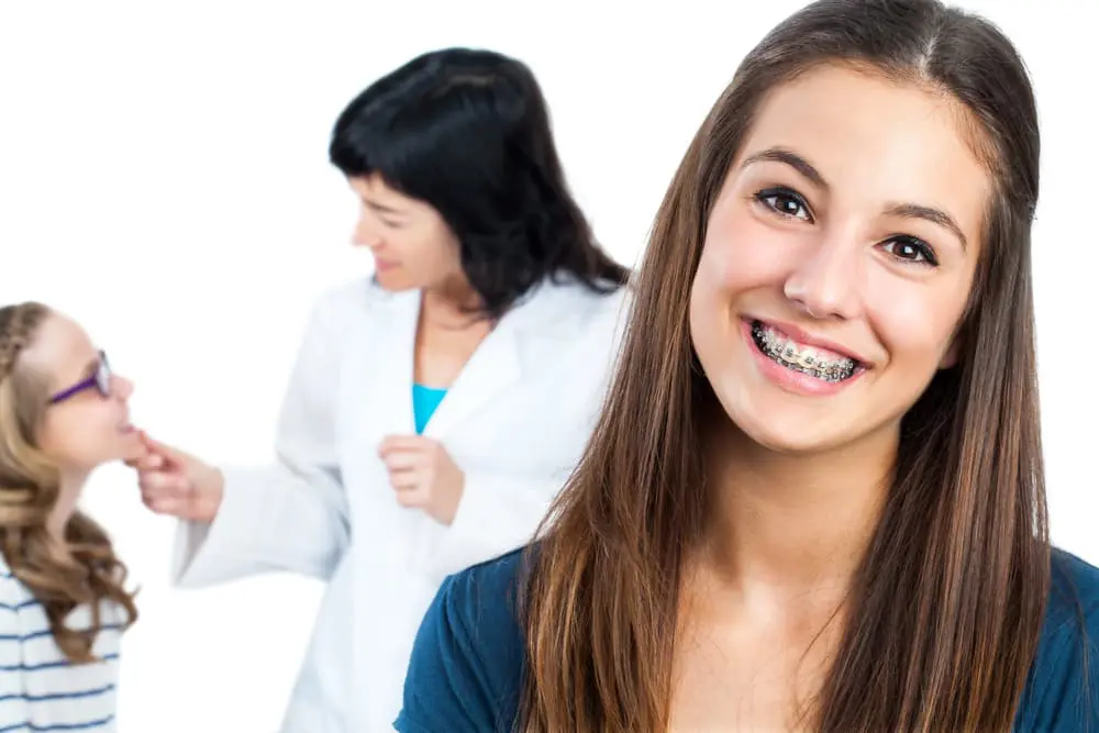 Best Orthodontics A smiling young woman with braces is in focus. In the background, a person who appears to be a dentist or orthodontist examines another young person. Chauncey Orthodontics in Henderson, NV can provide braces cost information.