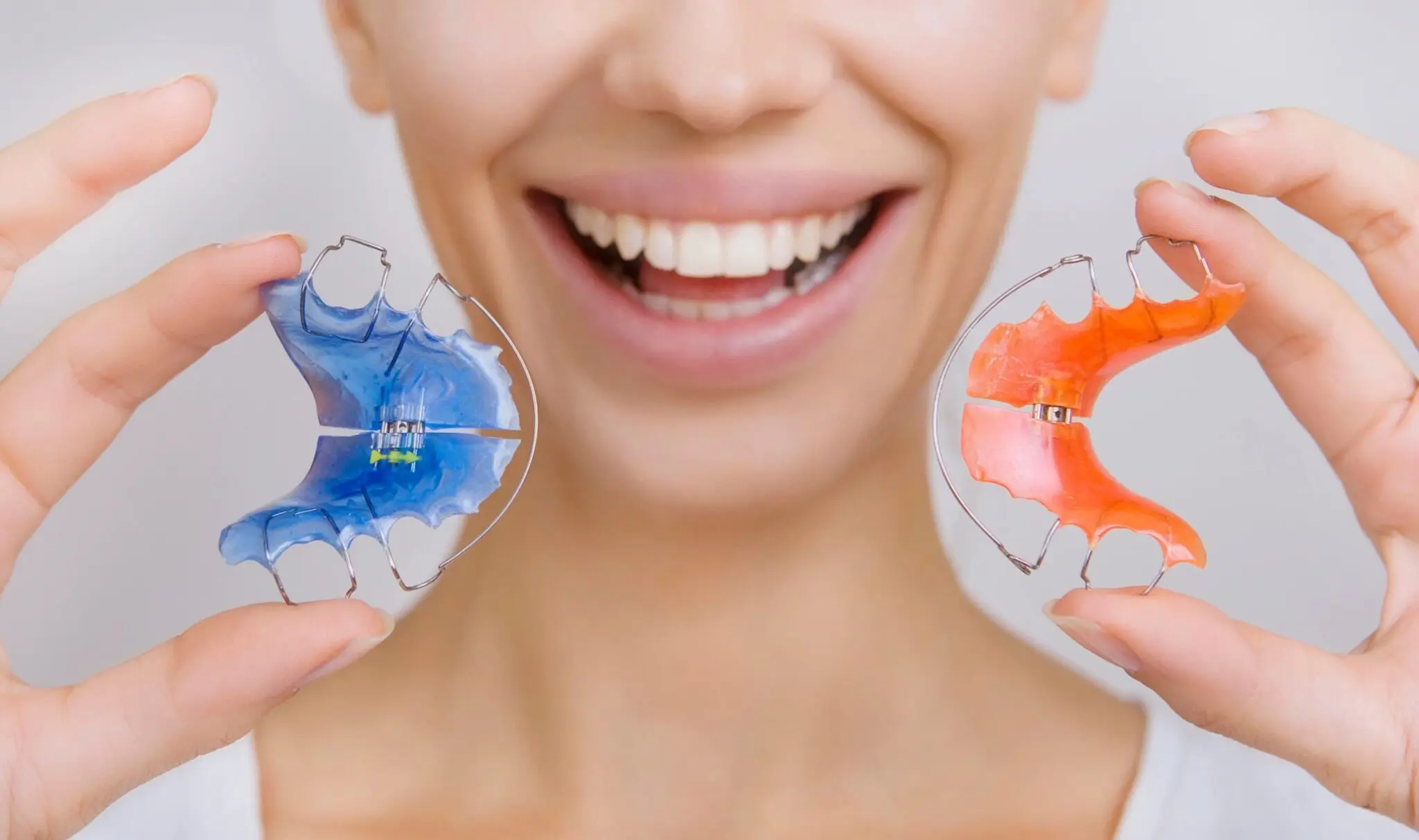 A person smiling and holding two orthodontic retainers, one blue and one orange, close to their mouth.