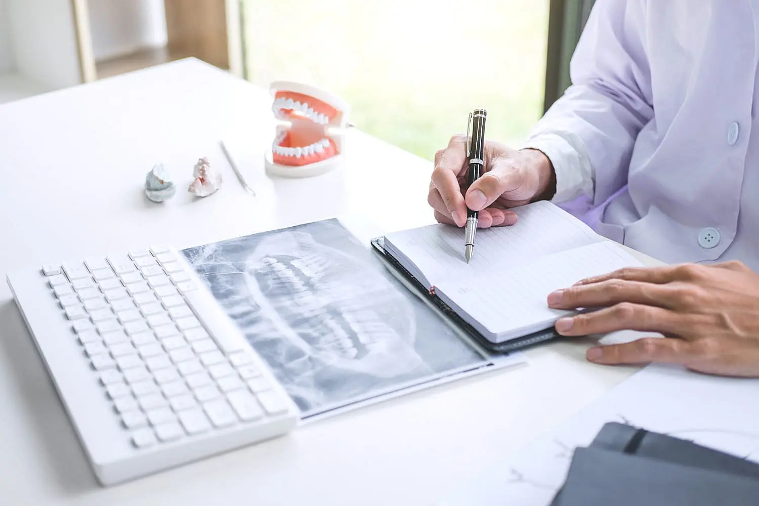 A person in a white coat writes in a notebook at a desk with a dental X-ray, likely reviewing orthodontic insurance plans in Henderson, NV.