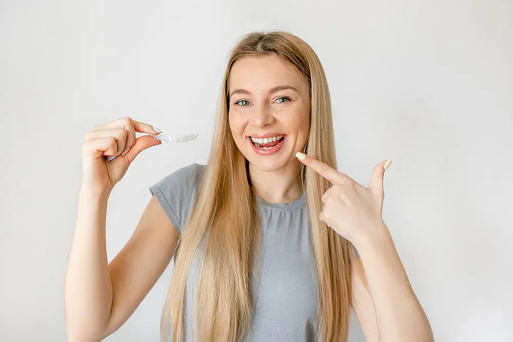 A woman with long blonde hair smiles while pointing to a clear dental aligner, showcasing the benefits of an Invisalign payment plan at Chauncey Orthodontics operates in Henderson, NV.