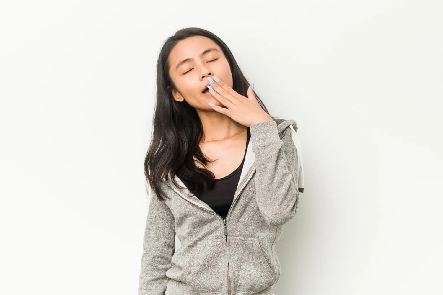 A woman in a grey hoodie is standing against a plain background, yawning with her eyes closed and covering her mouth with one hand cause mouth breathing issue in Henderson, NV.