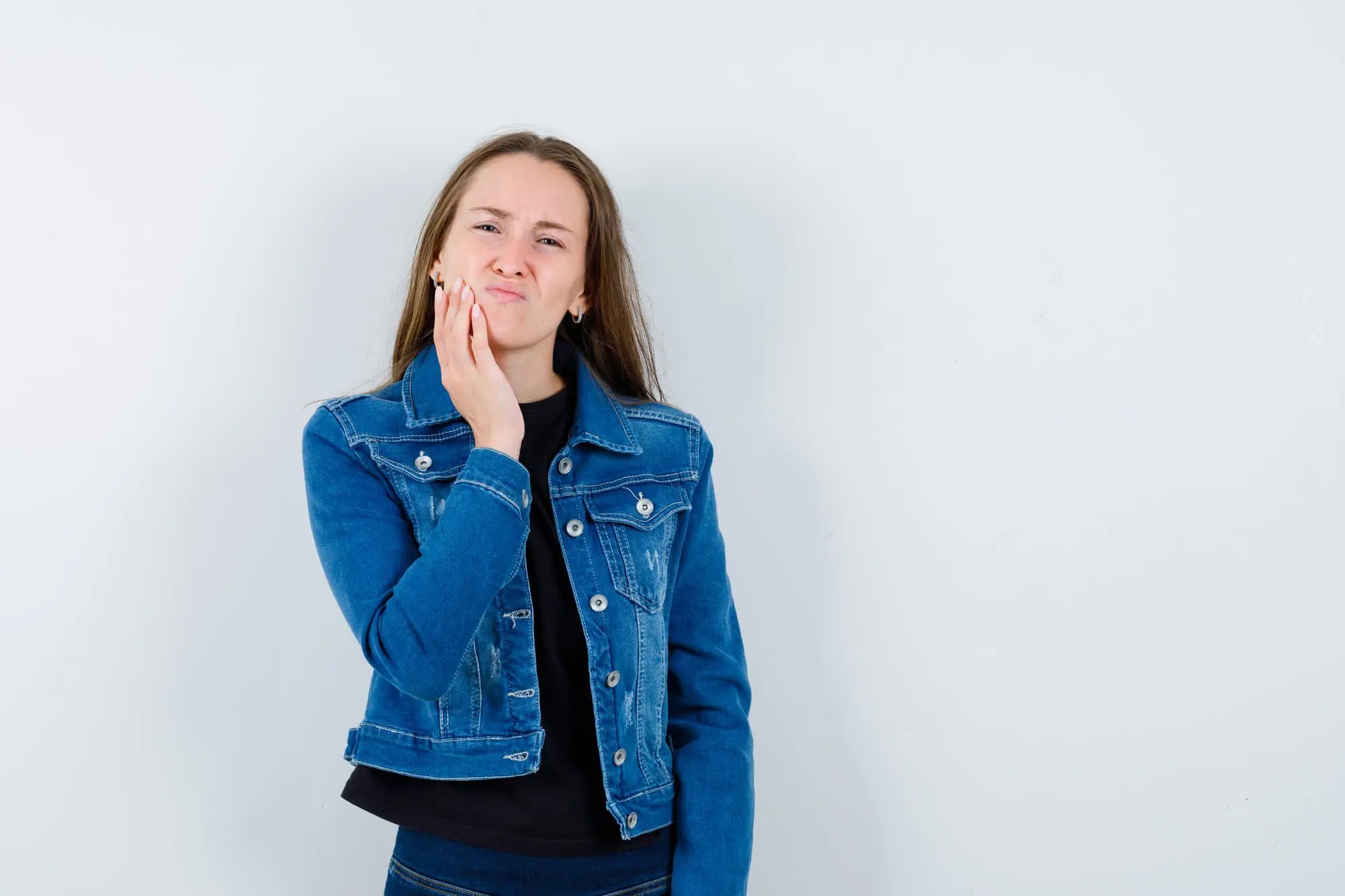 A woman in a denim jacket touches her cheek and looks uncomfortable, possibly experiencing underbite tooth pain, standing at Chauncey Orthodontics in Henderson, NV.