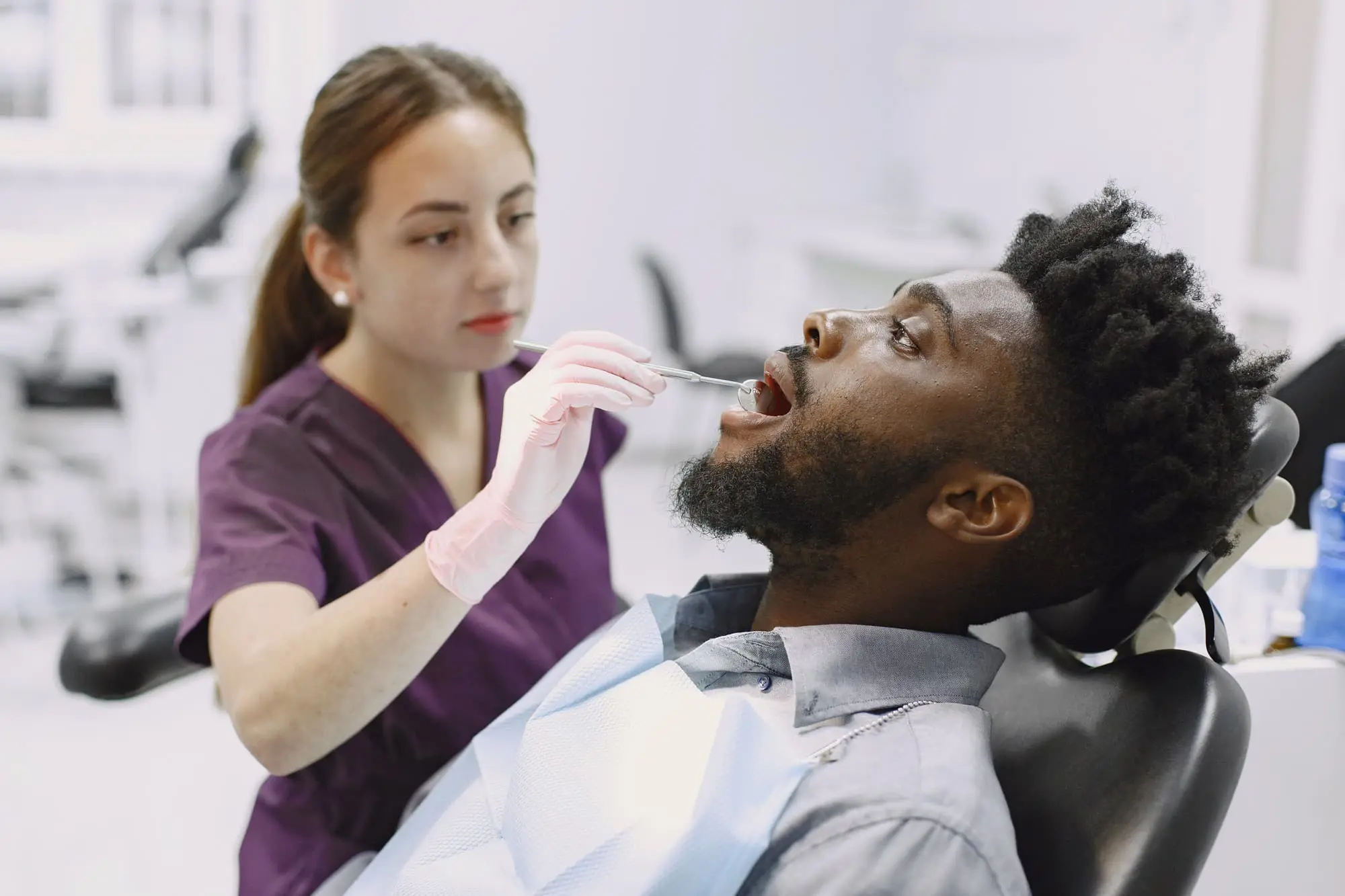 A dental professional examines a patient's mouth with a dental tool in a clinic setting. The patient is seated in a dental chair and wearing a bib.