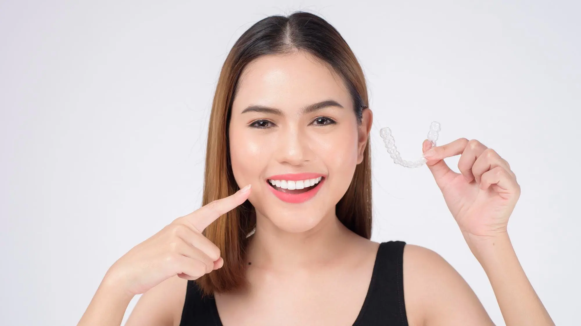 Woman smiling and holding a clear dental aligner, pointing to her teeth, representing the seamless Invisalign Payment Plan available at Chauncey Orthodontics operates in Henderson, NV.