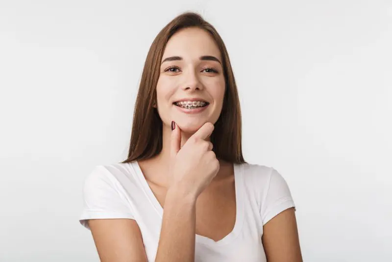 Young woman with long brown hair and metal braces smiles, wearing a white t-shirt, standing against a plain white background with her hand touching her chin at Chauncey Orthodontics in Henderson, NV.