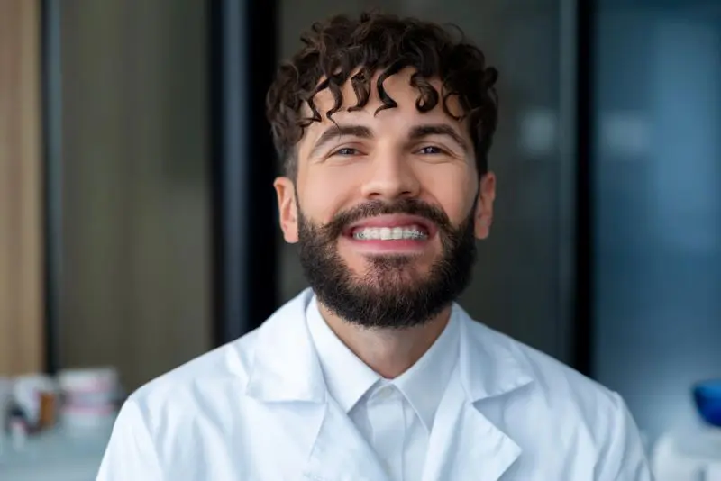 A person with ceramic brace and curly hair, wearing a white lab coat, smiles at the camera in an indoor setting in Henderson, NV.