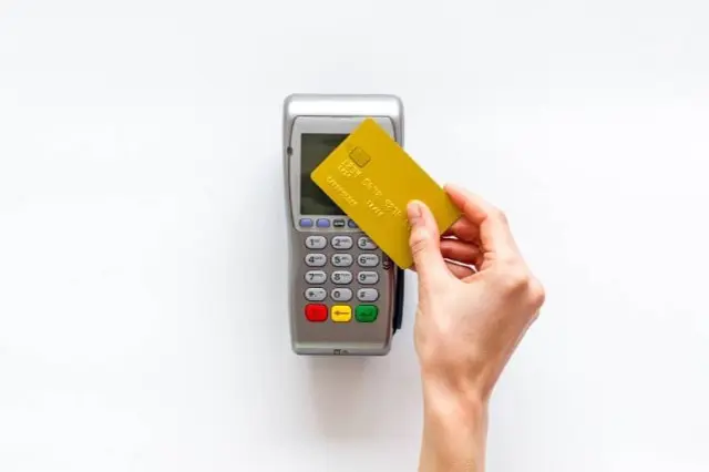 A hand holds a yellow credit card over a payment terminal with buttons and a display screen, ready to settle an braces and orthodontic insurance charge in Henderson, NV all set against a white background.
