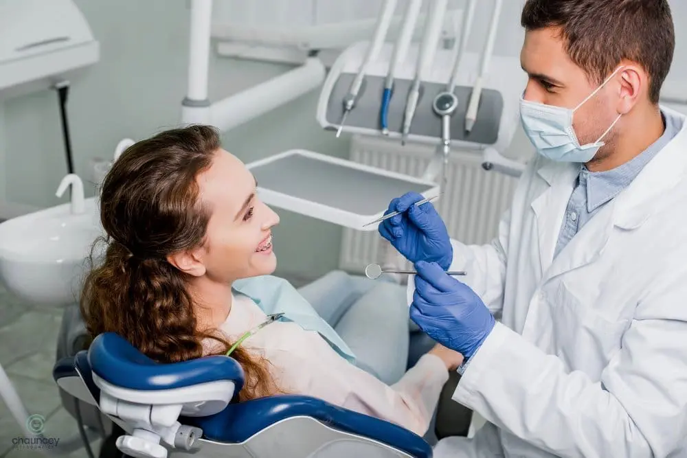 Orthodontist consulting a young girl about how to fix overbite in Henderson, NV with early intervention treatment. A dentist wearing a mask and gloves examines a smiling female patient in a dental chair, holding dental tools in a clinic setting.