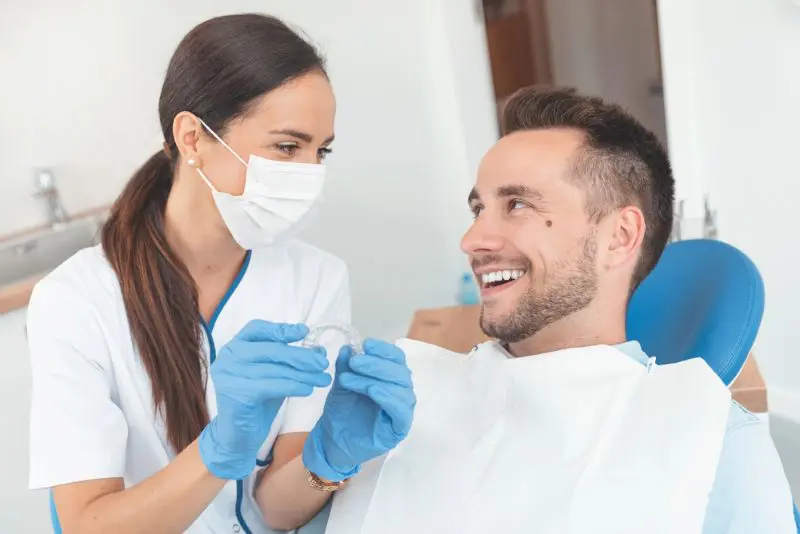 A dental professional wearing gloves and a mask shows a clear aligner to a smiling patient seated in a dental chair feel benefiting from orthodontic insurance plans at Chauncey Orthodontics in Henderson, NV.